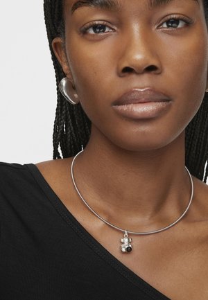 Collar choker hecho de metal plateado con un charm de oso que presenta acentos en blanco y negro. Modelo lleva una blusa negra con escote en V.