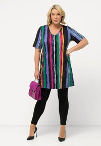 Túnica a rayas multicolores con escote en V, que presenta rayas moradas, rosas, verdes y negras, combinada con leggins negros y un bolso morado.