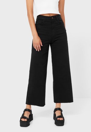 Czarne szerokie spodnie z bawełny, z wysokim stanem, przednimi kieszeniami i surowym brzegiem. Dobrane do nich masywne czarne sandały na platformie.