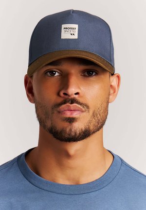 Man met een getrimde baard, die een blauw-bruine honkbalpet draagt met een 'PROTEST SINCE '93'-badge en een blauw shirt met ronde hals, op een neutrale achtergrond.