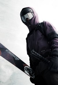 Skier wearing black helmet, goggles, and purple jacket holding Nordica skis and ski poles against snowy background.