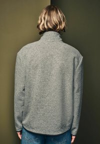 Sivý pulóver s vysokým golierom, vyrobený z mäkkej tkaniny, s uvoľneným strihom a spustenými ramenami, v kombinácii s modrými džínsami.