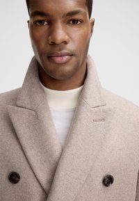 Uomo che indossa un cappotto beige di lana doppiopetto sopra un maglione bianco a collo alto a coste, su uno sfondo neutro.