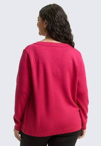 Eine Frau mit dunklem, lockigem Haar trägt ein lockeres, langärmliges, leuchtend pinkes Oberteil und schwarze Hosen und steht mit dem Rücken zu einem schlichten, hellen Hintergrund.