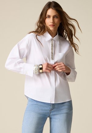 Frau trägt ein weißes Hemd mit Knöpfen, verziert mit silbernen Streifen und Armbändern, kombiniert mit hellblauen Jeans vor einem neutralen Hintergrund.