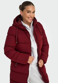 Abrigo acolchado rojo con capucha, que presenta un cierre frontal y un diseño de acolchado. Llevado sobre una sudadera gris con un acabado suave y brillante.