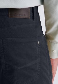 Black corduroy trousers with vertical ribbing, featuring a brown leather tag with embossed text and a side pocket detail. Light-colored shirt visible.