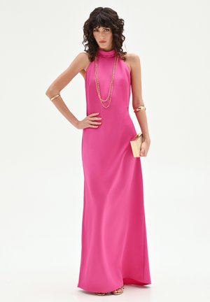 Femme aux cheveux bouclés et foncés portant une robe rose longue sans manches, un collier en chaîne dorée, des bracelets, et tenant une petite pochette en bois.