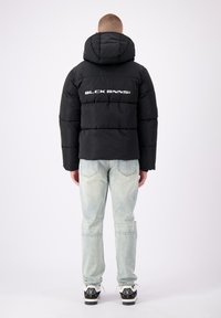 Zwarte pufferjas met capuchon, voorzien van horizontaal gestikte secties en een wit logo op de achterkant. Gecombineerd met lichtblauwe spijkerbroeken en sneakers.