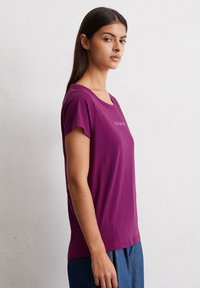 Paarse T-shirt met korte mouwen, een ruime hals en een losse pasvorm, met een kleine tekstdetail op de voorkant. Zacht stof met een gladde textuur.