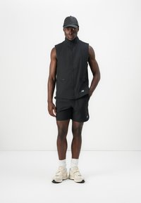 Crni bezrukavni prsluk s visokim ovratnikom, dva džepa sa zatvaračem, upareni s crnim kratkim hlačama. Outfit je upotpunjen kremastim tenisicama i crnom kapom.