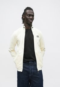 Lysecreme lynlåstrøje med lodret ribbet tekstur, sorte accenter på kraven, kombineret med en sort t-shirt og mørkeblå jeans.