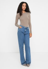 Kvinne med langt krøllete hår iført en tettsittende taupe langermet topp, løse blå jeans og spisse taupe sko, stående med en hånd i lommen.