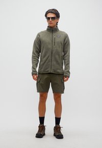 Άνδρας που φοράει φλις με φερμουάρ σε ελιά πράσινο, cargo σορτς, μποτάκια πεζοπορίας, μαύρες κάλτσες και γυαλιά ηλίου, στέκεται μπροστά σε λευκό φόντο.