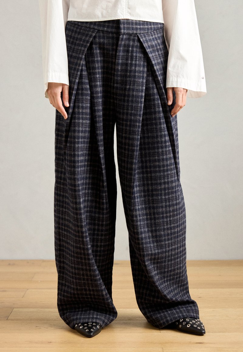 Mornarske karirane hlače širokih nogavica od teksturiranog materijala s plitkim nabora na struku, uparene s ukrašenim crnim cipelama sa srebrnim detaljima.