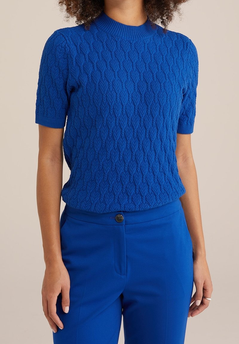 Femme portant un haut en maille bleue texturée à manches courtes et un pantalon bleu ajusté avec fermeture par un bouton sur un fond uni.