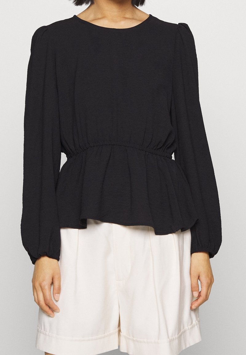 Femme portant un haut péplum noir à manches longues et un short blanc ample arrivant au genou sur un fond uni.