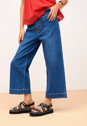 Femme portant un jean large court bleu, des sandales tressées noires et blanches, un haut rouge avec dentelle, et des bracelets au poignet.