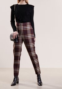 Schwarzes, geripptes Langarmshirt mit Rüschendetails, kombiniert mit hochtaillierten Hosen in Pflaumen- und Brauntönen mit Karomuster und schwarzen Stiefeletten, während eine kleine Tasche gehalten wird.