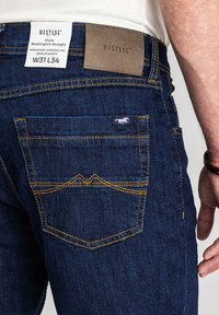 Ciemnoniebieskie dżinsy z denimu o średnim stanie i prostych nogawkach. Charakteryzują się żółtym przeszyciem, skórzaną naszywką oraz małym logo marki na kieszeni.