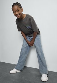 Enfant souriant avec des cheveux tressés portant un t-shirt gris foncé, un jean bleu clair et des baskets blanches, penché en avant avec les mains jointes.