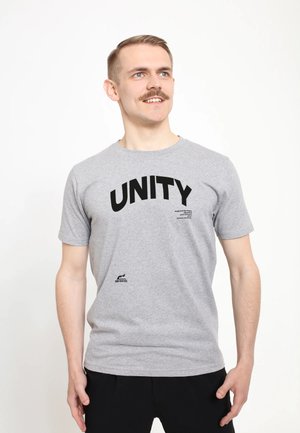 Mężczyzna z wąsami w jasnoszarej koszulce z dużym czarnym napisem "UNITY", stojący na białym tle, patrzący w górę i uśmiechający się.