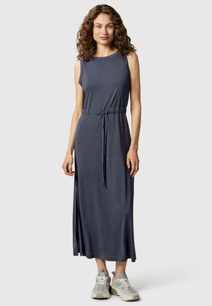 Frau mit lockigem Haar, die ein ärmelloses dunkelgraues Maxikleid mit Seitenschlitz und beige Sneakers trägt, stehend vor einfachem Hintergrund.
