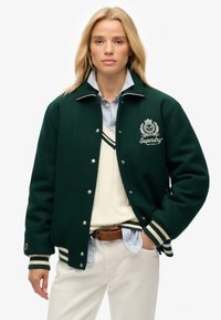 Groene wollen varsityjack met witte accenten, voorzien van gestreepte manchetten en kraag, geborduurd logo op de borst, gedragen over een crèmekleurige trui.