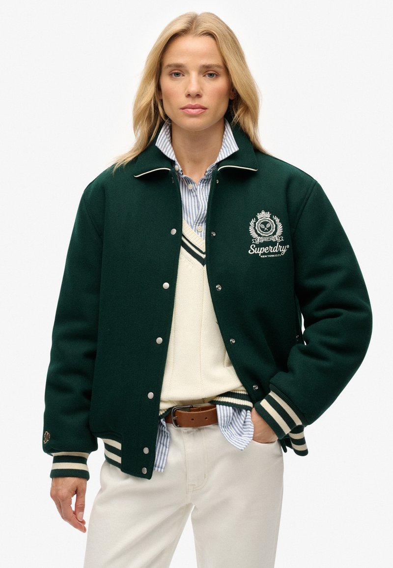 Groene wollen varsityjack met witte accenten, voorzien van gestreepte manchetten en kraag, geborduurd logo op de borst, gedragen over een crèmekleurige trui.