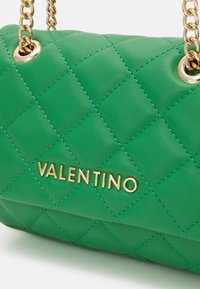 Zelená prošívaná kožená kabelka se zlatým kováním, vybavená řetízkovým páskem a vyraženým nápisem značky "VALENTINO" ve zlaté barvě.