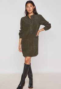 Abito di velluto a coste verde oliva con un collo henley bottonato, maniche lunghe, due tasche frontali e una vestibilità morbida, abbinato a stivali neri fino al ginocchio.