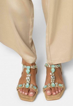 Pies con sandalias de punta cuadrada en beige adornadas con tiras de joyas azul claro y transparentes, combinadas con pantalones de seda beige.