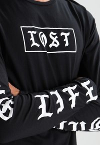 Long-sleeve black shirt with white graphic text. "LOST" in a rectangle on the front, "LIFE" running vertically on each sleeve. Soft fabric, relaxed fit.