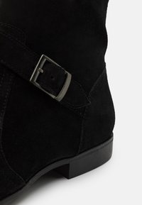 Svart mocka ankelboot med en silverfärgad spännsrem och låg staplad klack på en vit bakgrund.