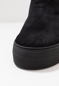 Černé semišové kotníkové boty s texturovanou gumovou podrážkou, kulatou špičkou a prošívanými detaily. Svršek má hladký, jemný povrch.