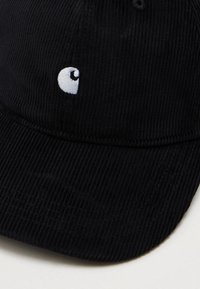 Gorra de béisbol de pana negra con logo bordado en blanco en el centro frontal, colocada sobre una superficie clara y lisa.