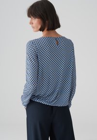 Blaue Bluse mit geometrischem Muster, leichter Stoff, lockere Passform und einem Schlüssellochdetail auf der Rückseite, kombiniert mit dunklen Hosen.