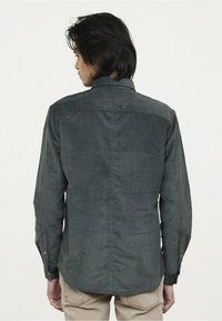 Chemise en velours côtelé vert foncé avec un col à boutons, des manches longues et une texture lisse. Présente des coutures visibles et un ourlet arrondi.