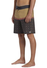 Uomo che indossa pantaloni da bagno multicolore con motivo geometrico, in piedi a piedi nudi su uno sfondo bianco.