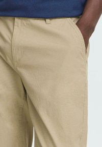 Beige cotton blend trousers with a flat front, side pockets, and a relaxed fit, featuring subtle texture and minimal stitching details.
