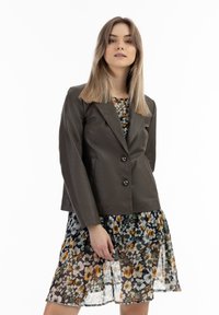 Bruin leren blazer met een getailleerd ontwerp, twee grote knopen, gelapte revers; gedragen over een bloemen, gelaagde, doorschijnende jurk in verschillende kleuren.