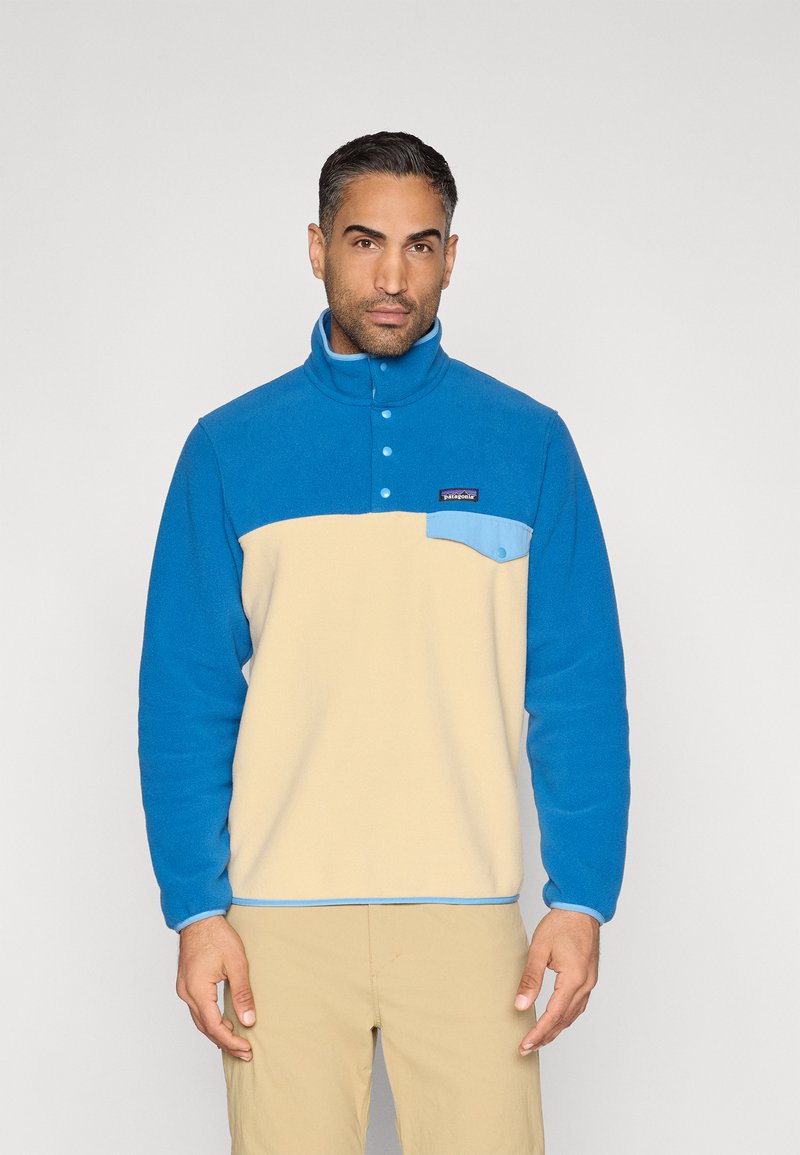 Φλις πουλόβερ σε μπλε και μπεζ χρώμα, με ψηλό γιακά, εμπρός κουμπώματα και τσέπη στο στήθος με ανοιχτό μπλε λεπτομέρεια. Μαλακή υφή.