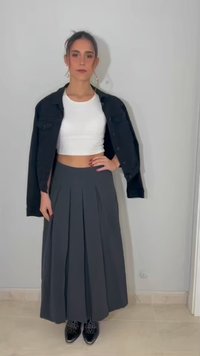 Femme debout contre un mur clair portant un crop top blanc, une veste noire posée sur les épaules, une longue jupe plissée grise et des chaussures noires décorées.