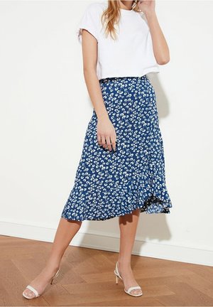 Žena v bílém topu s krátkými rukávy, modré midi sukni s bílým květinovým vzorem a bílých sandálech na podpatku stojí na dřevěné podlaze.