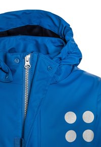 Blå jacka med silverdragkedja och fyra runda reflexprickar på bröstet, med hög krage och fast huva.