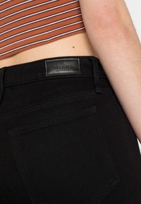 As calças de ganga pretas possuem um emblema de pele com "HOLLISTER CALIFORNIA" em relevo. O tecido é suave, com detalhes de costura mínimos.