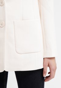 Light cream blazer made of textured fabric, featuring a single chest pocket, five lightly colored buttons, and a tailored fit.