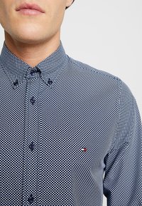 Uomo che indossa una camicia button-down blu navy con pois bianchi e un piccolo logo rosso, bianco e blu sul petto.