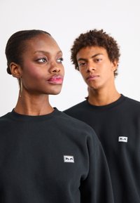 Mustad mustrid, ribatud kaelusega, millel on väike logoäpp vasakul rinnal. Kangas näib olevat pehme ja lõige on mugav.