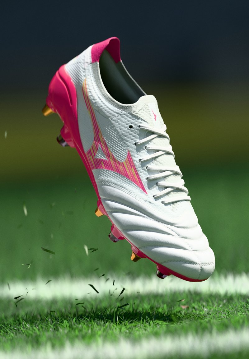 White and pink soccer cleat with gold studs in mid-air above grass on a sports field near a white line.